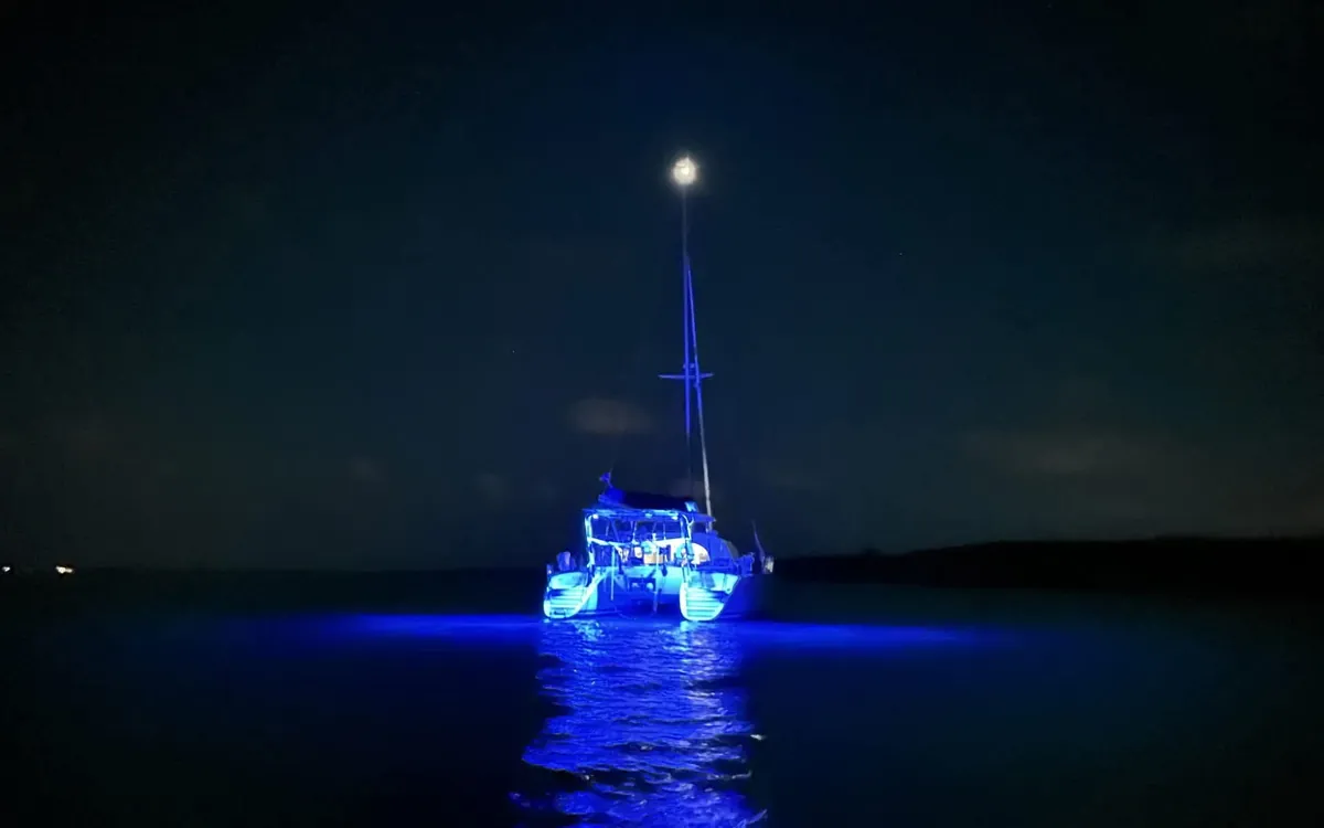 Majik at anchor after dark, hulls lit blue by her underwater lights, the moon on the masthead